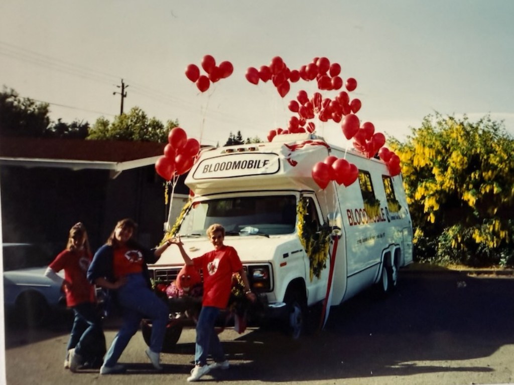 An early Bloodmobile decorated with red balloons and surrounded by staff